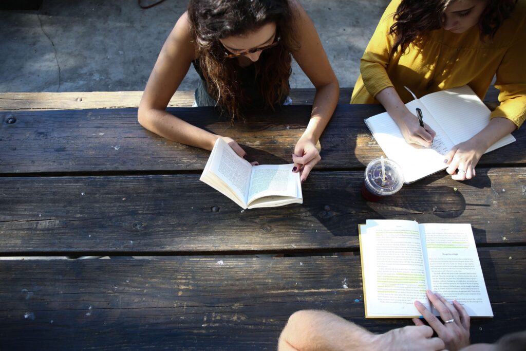 Two people are sitting at a wooden table outdoors, engrossed in studying and taking notes. One writes in a notebook while the other reads dystopian books for teens. An iced drink is placed nearby on the table, its condensation glistening in the warm sunlight.