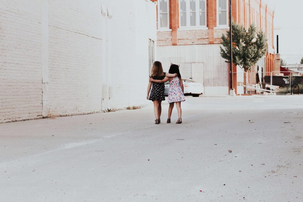 Two girls in summer dresses walk arm-in-arm down an empty street lined with brick buildings, engaged in cheerful chatter about their favorite get-to-know-you games for kids. One wears a black dress with white spots; the other wears a floral dress. The atmosphere is sunny and peaceful.