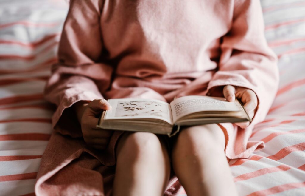 A child wearing a pink outfit sits on a bed with striped pink and white sheets, absorbed in reading one of the Boxcar Children books. The focus is on the hands and the open book, crafting a cozy and peaceful scene.