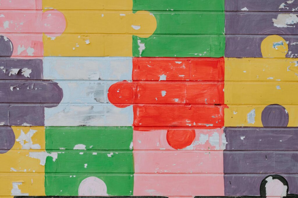 A colorful mural of interlocking puzzle pieces on a wooden surface features shades of yellow, green, red, pink, purple, and white. The weathered and peeling paint adds texture to this artistic homage to educational puzzles for kids.