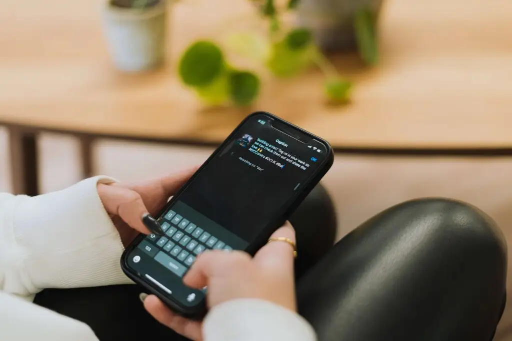 A person in a white sweater and black pants is seated, absorbed in typing on a smartphone. The screen displays a dark-themed messaging app—a digital hub for sharing moments or seeking answers to why people post on social media. In the blurred background, a small plant graces the wooden table.