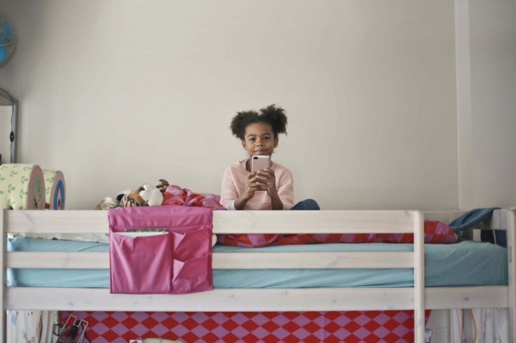 A child with curly hair sits on a bunk bed, smiling and holding a kid-friendly dumb phone. The bed is unmade with colorful sheets and a pink pocket organizer on the side. The room has a neutral color scheme and a decorated wall behind.