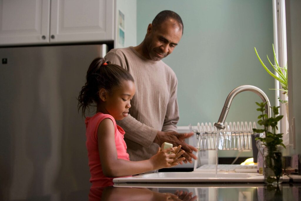 A man and a young girl, learning how to talk so kids will listen, wash their hands together at the kitchen sink. The man wears a beige shirt, the girl a pink sleeveless top. Sunlight streams in, with a drying rack and plant nearby.