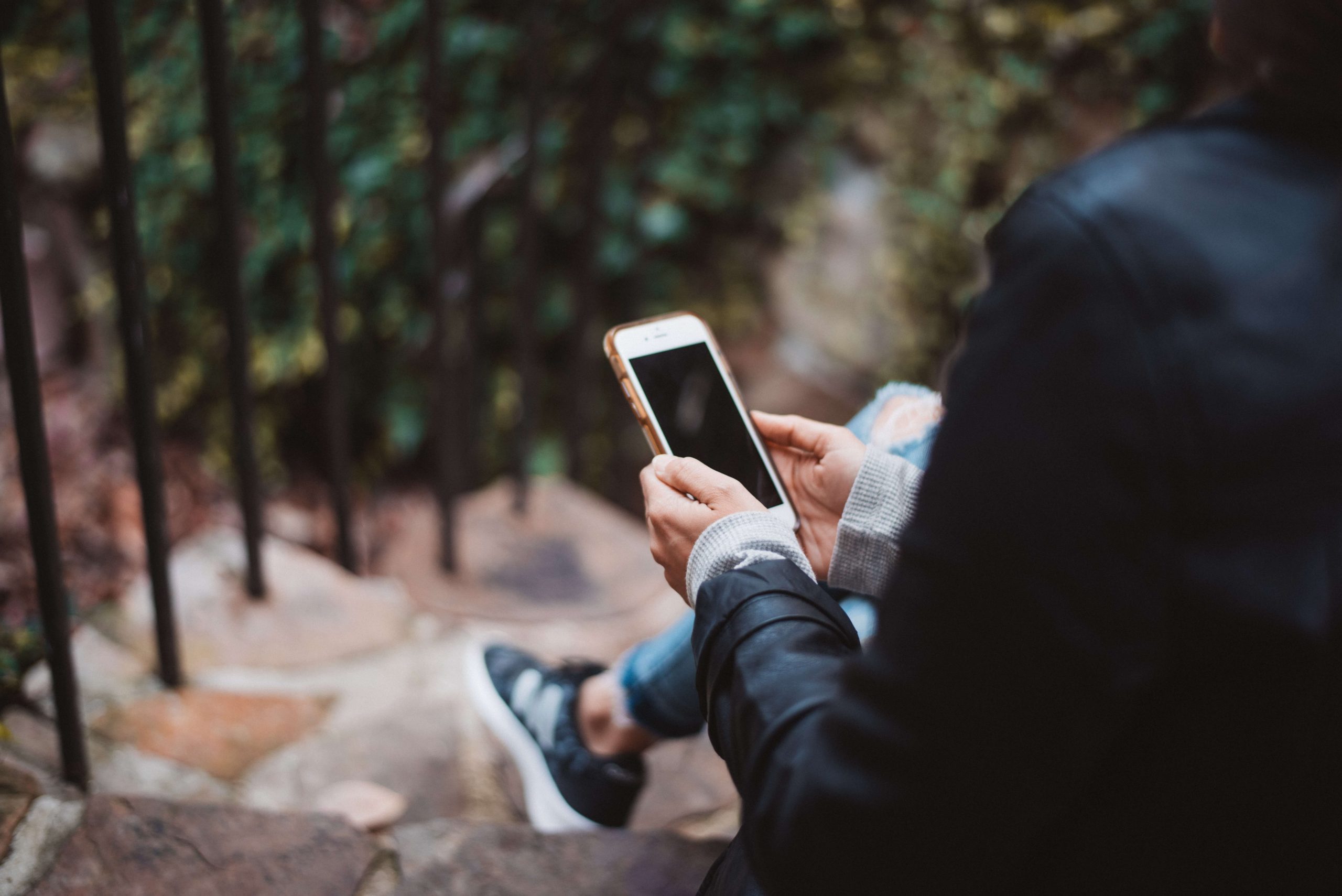 A person in a black jacket and blue jeans sits on outdoor stone steps, engrossed in their smartphone. As the blurred foliage and black railing create a serene background, they remain unaware of the many types of cyberbullying that unfold across screens worldwide.