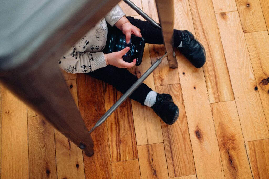 A child sits on a wooden floor, partially hidden under a chair, playing with a smartphone. It's always a debate about the good age to give kids a phone. Wearing black shoes and a white sweatshirt with patterns, the child's curiosity with technology is unmistakable.