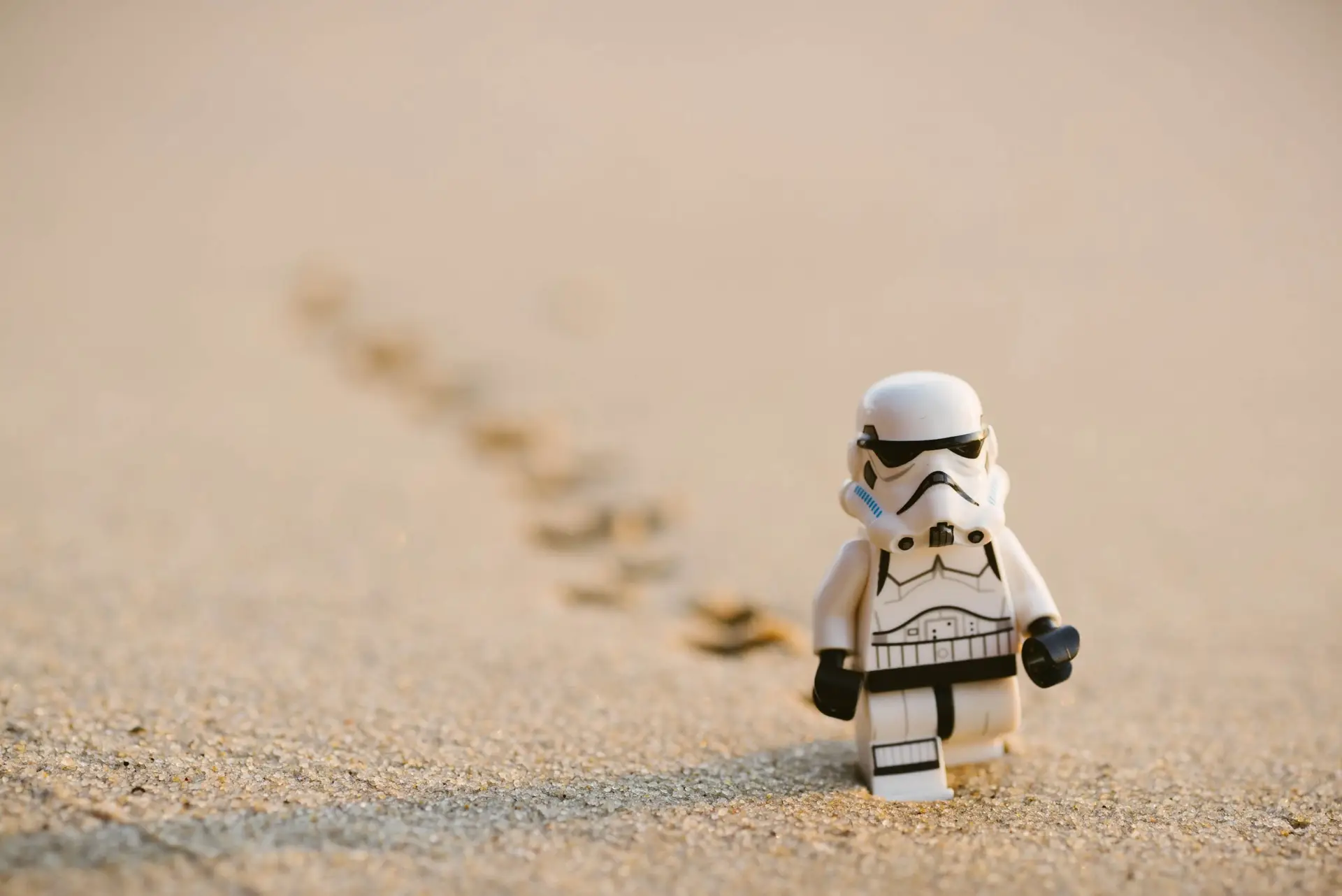 A small toy figure dressed as a stormtrooper marches across sandy terrain near a Star Wars-themed food stall, leaving a trail of footprints behind. The softly blurred background highlights the solitude and simplicity of this charming scene.