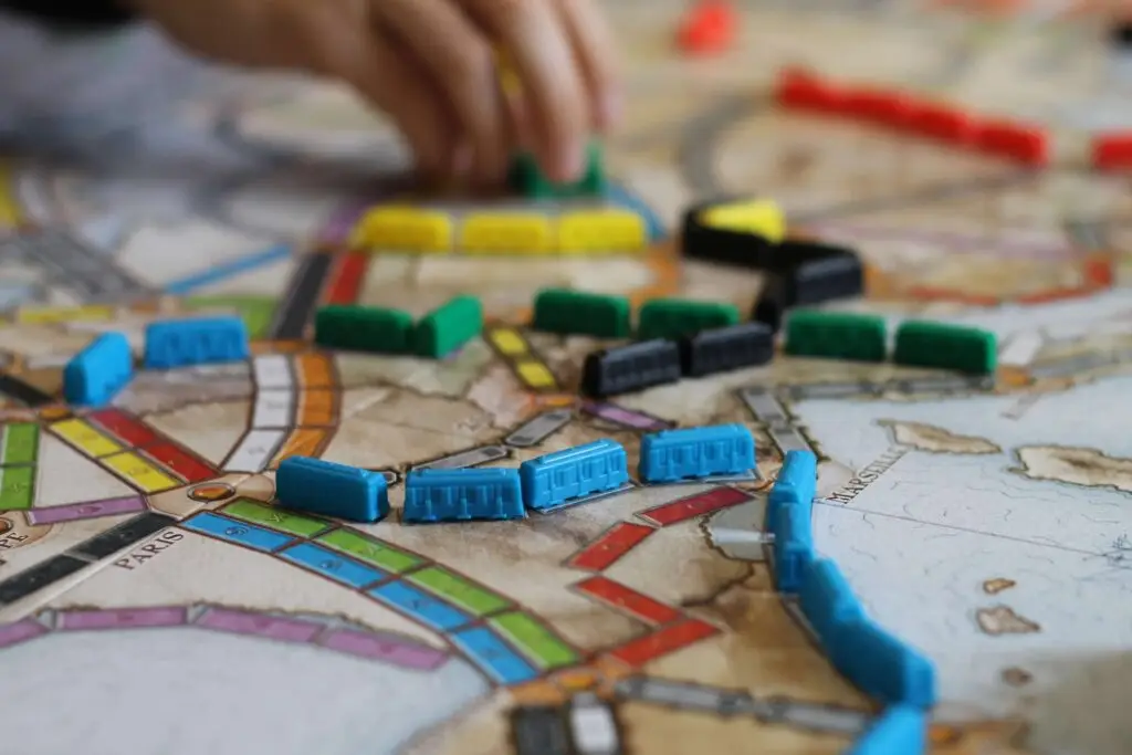 A hand arranges colorful train pieces on a game board map, reminiscent of math board games for kids. The board shows various cities connected by tracks in red, blue, green, yellow, and black. The image suggests an engaging board game in progress.