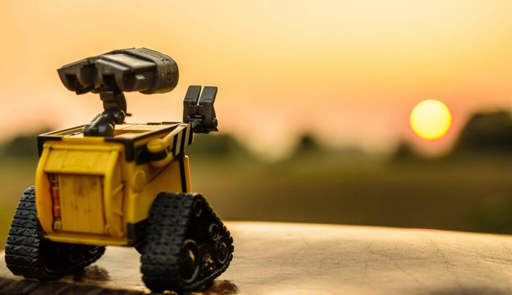 A small yellow robot with tank-like wheels gazes at a scenic, glowing sunset, much like a scene straight out of beloved robot movies for kids, with the sun setting on the horizon and a blurred landscape in the background.