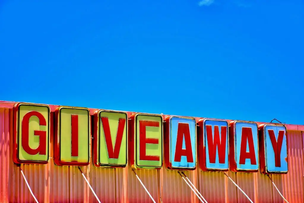 A colorful sign with large, bold letters spelling GIVEAWAY stands against a bright blue sky. The letters are in individual, framed blocks on a corrugated metal backdrop, evoking a vintage aesthetic reminiscent of nostalgic back-to-school days.