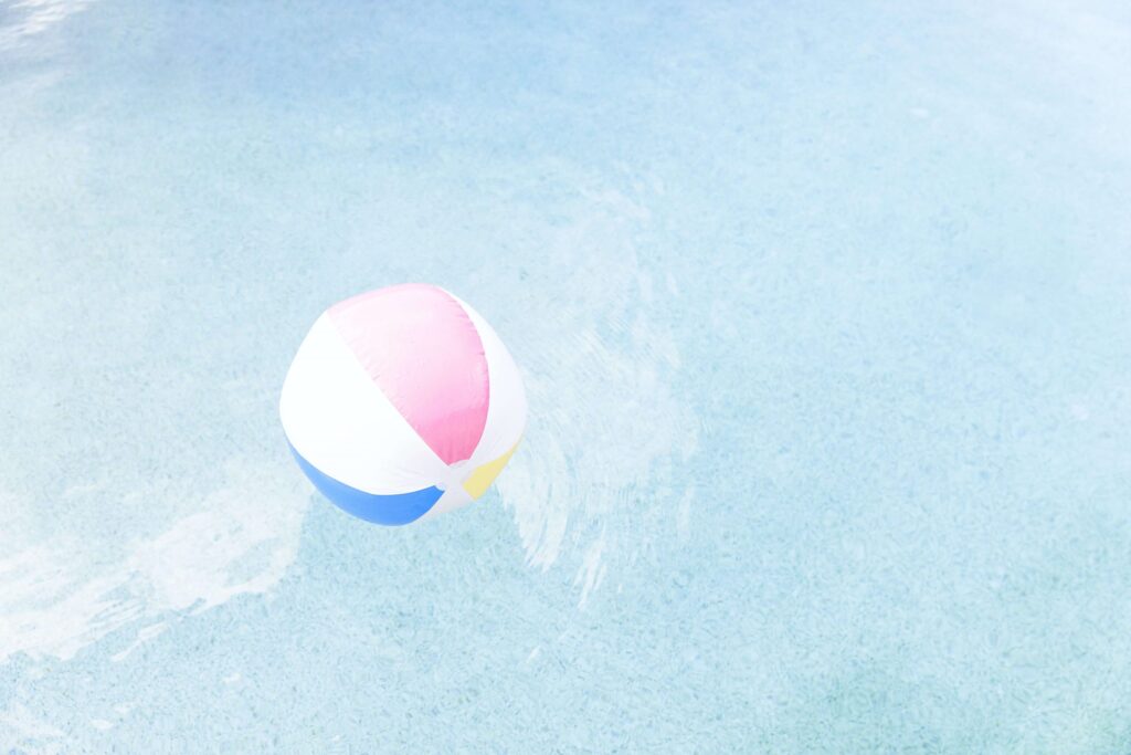 A colorful beach ball floats on the surface of a clear blue swimming pool, perfect for pool games for kids. It features segments of pink, white, yellow, and blue, creating gentle ripples in the water.