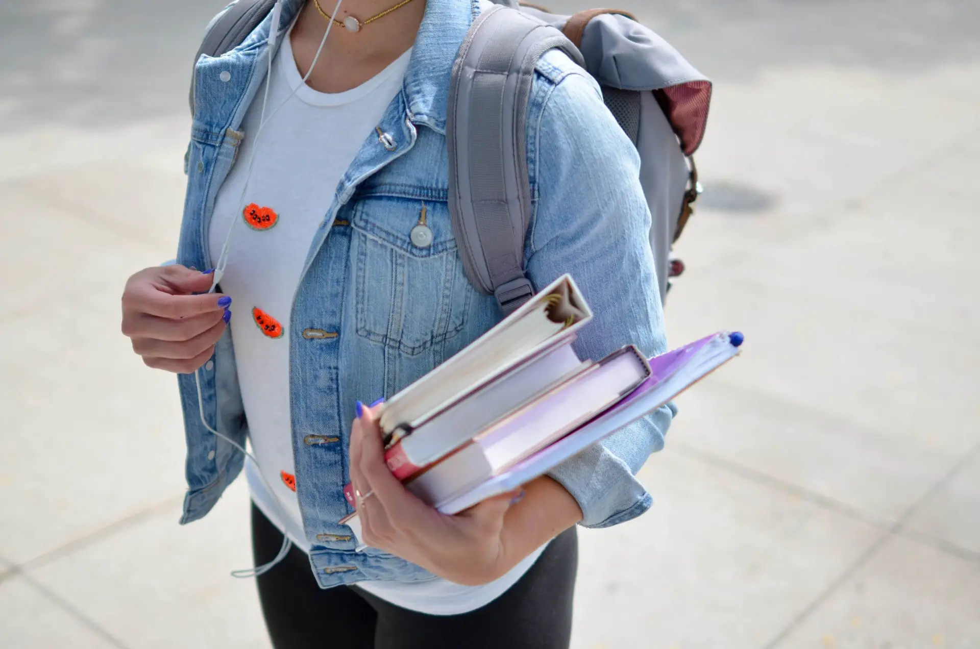 A person wearing a denim jacket and backpack, likely filled with show and tell ideas for kids, holds a stack of books while strolling down the sidewalk. Earbuds dangle from their ears, complementing a white shirt adorned with vibrant orange patches.