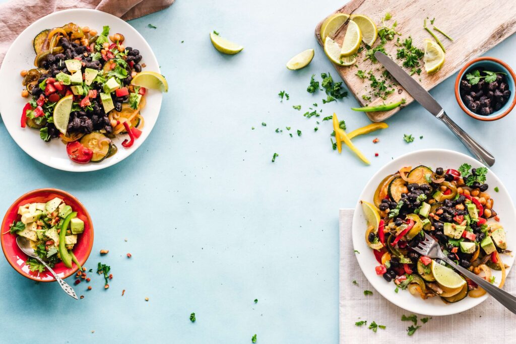 Two plates of vibrant salad featuring black beans, diced avocado, bell peppers, and lime wedges rest on a light blue surface—perfect for those seeking gluten-free, dairy-free recipes. A small bowl with similar ingredients and a cutting board with fresh herbs and lime slices completes the scene.