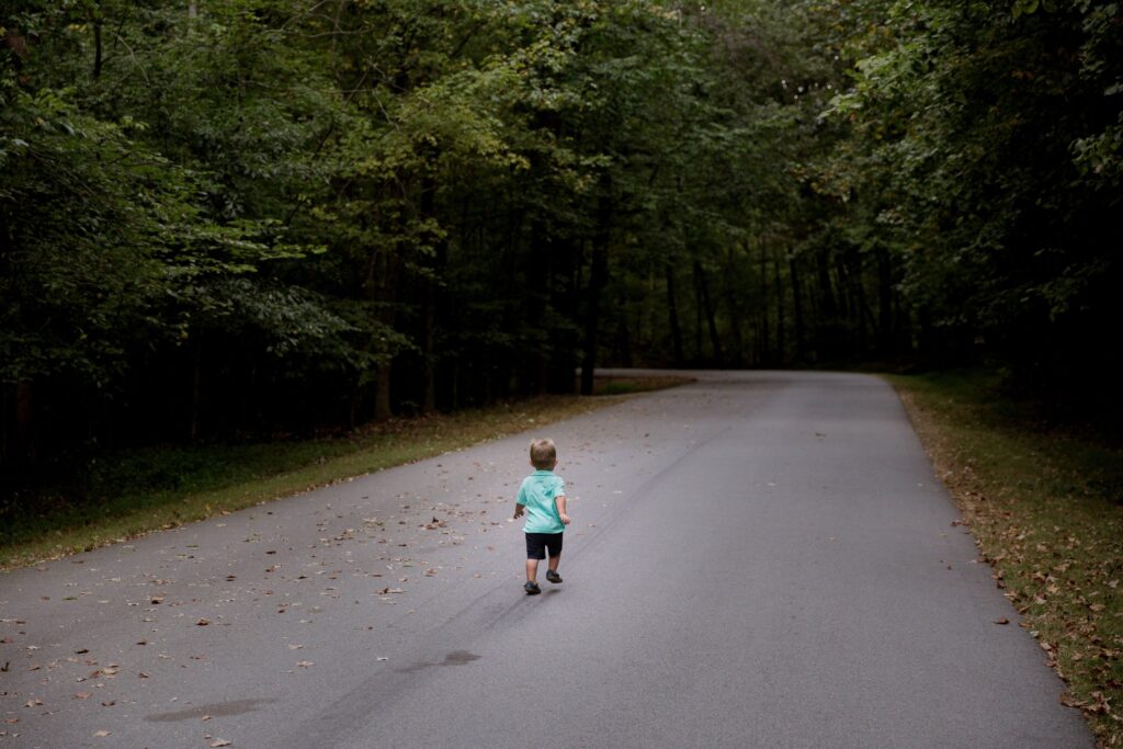 A young child in a green shirt walks along a winding, tree-lined road, with fallen leaves scattered on the pavement. As the dense forest ensures a serene atmosphere, one wonders how much time kids should spend on phones amidst nature's beauty.