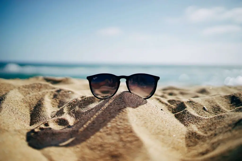 A pair of black sunglasses is partially buried in the sandy beach with the ocean and blue sky in the background, like hidden summer riddles for kids waiting to be discovered. The focus is on the sunglasses, creating a relaxed, sunny atmosphere.