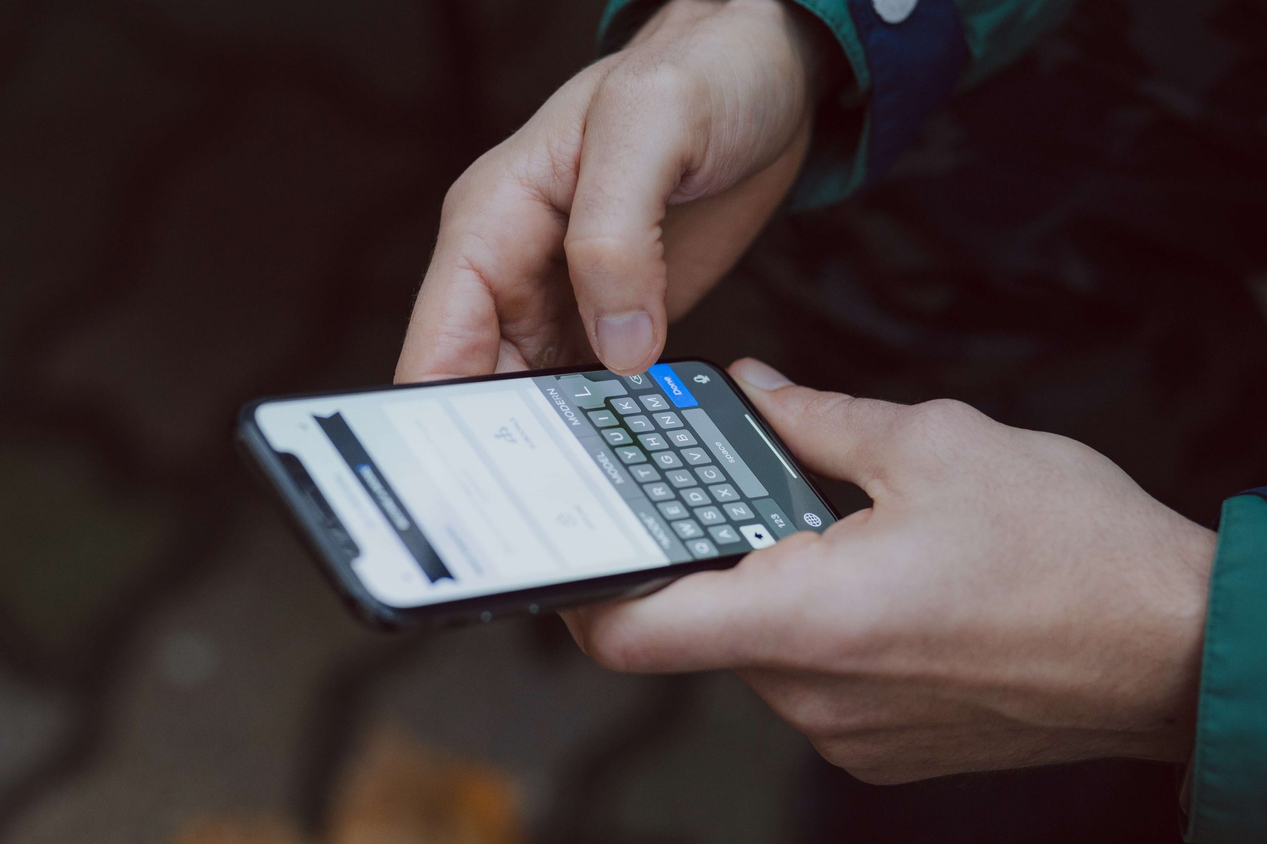 A person wearing a dark jacket is holding a smartphone, designed as an emergency phone for kids, with both hands, typing on the on-screen keyboard. The background is out of focus, revealing stone pavement while the phone screen displays a conversation with text input.