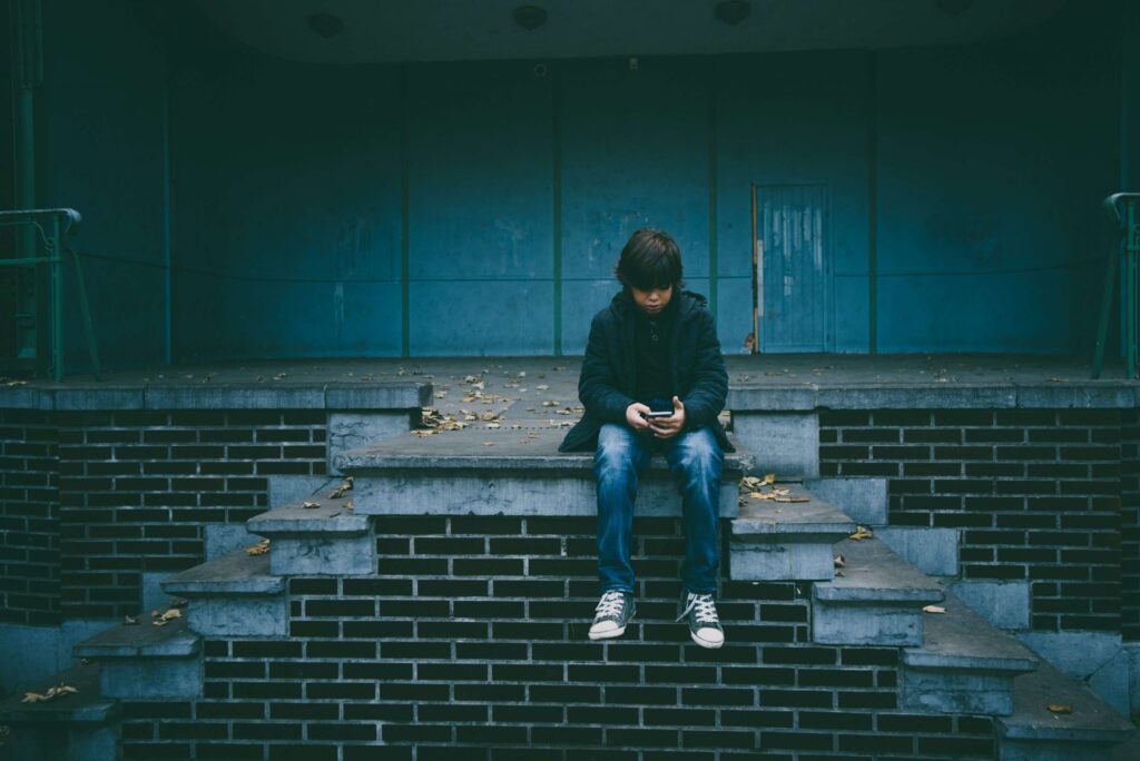 A solitary person sits on concrete and brick steps, absorbed in a smartphone, pondering how social media affects social skills. Fallen leaves surround them, and the background is a dark, empty stage. The mood is calm and reflective.