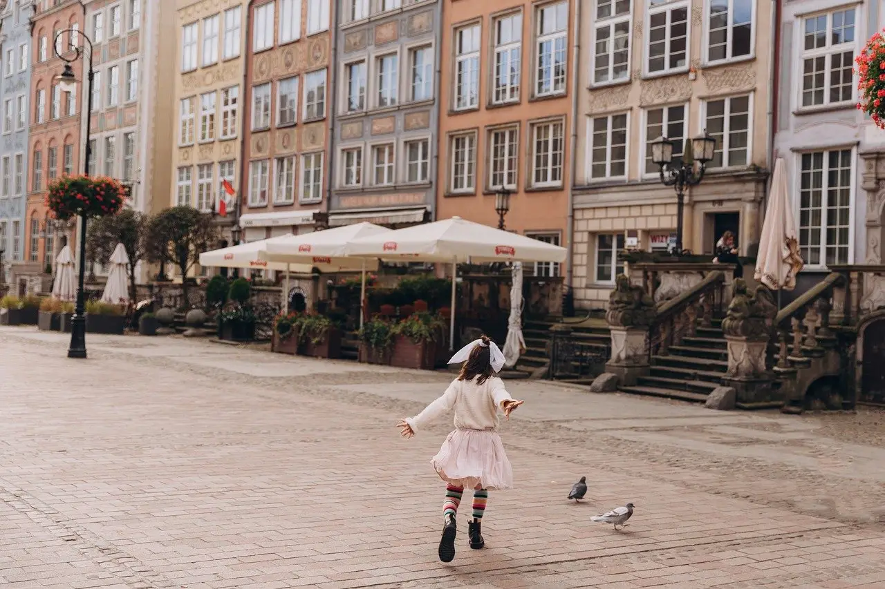 A child in a pink skirt and striped stockings bursts with energy, turning pigeon-chasing into one of the spontaneous ADHD activities for kids. They navigate the cobblestone street lined with colorful historic buildings and outdoor cafes, while bright flowers hang from poles on a cloudy day.