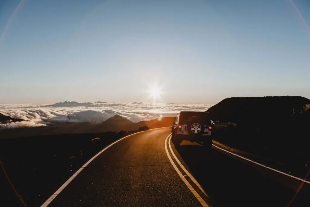 A car is parked on a winding mountain road during sunset, setting the scene for travel games for kids as the journey unfolds. The road leads toward a horizon filled with clouds and a glowing sun, creating a serene and picturesque landscape.