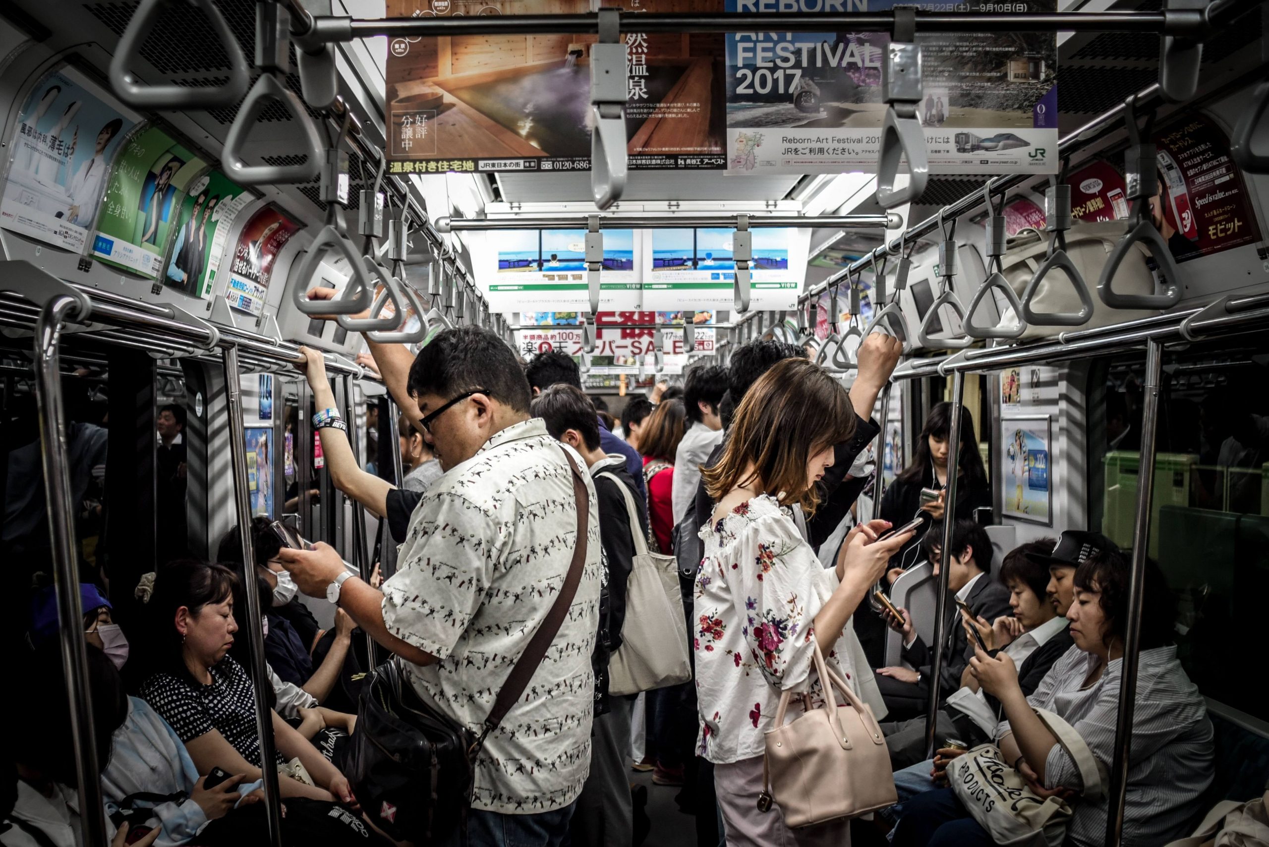 The crowded subway train buzzes with passengers, most immersed in their smartphones, reflecting on how many people are addicted to social media. Overhead handles dangle from the ceiling, ads peek above, and casually dressed passengers fill the bright car.