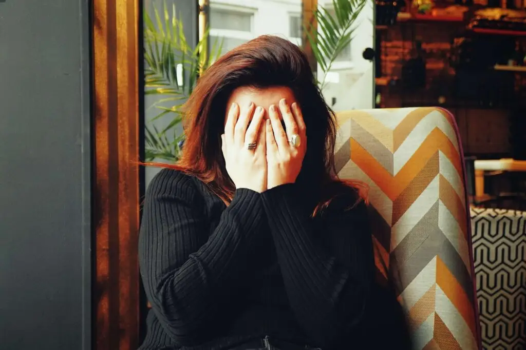 A person with long hair sits in a chair, covering their face with their hands, perhaps feeling easily embarrassed. They wear a black sweater and several rings. The chair has a zigzag pattern, and plants and a window fill the background.