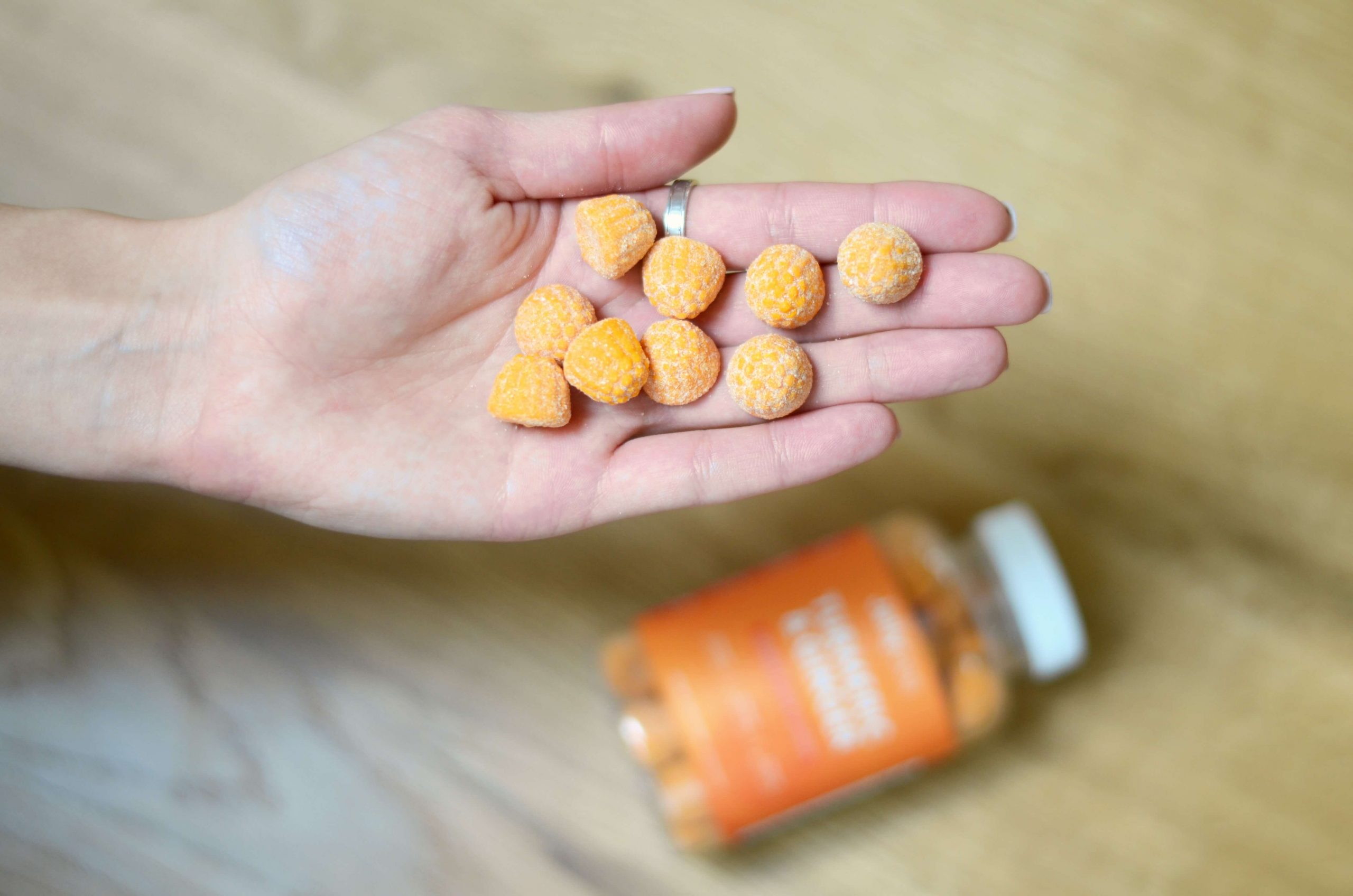 A hand holds several small, round orange gummies, each a burst of focus-boosting vitamins to help kids concentrate better. In the background, a bottle of vitamin gummies rests on a wooden surface.