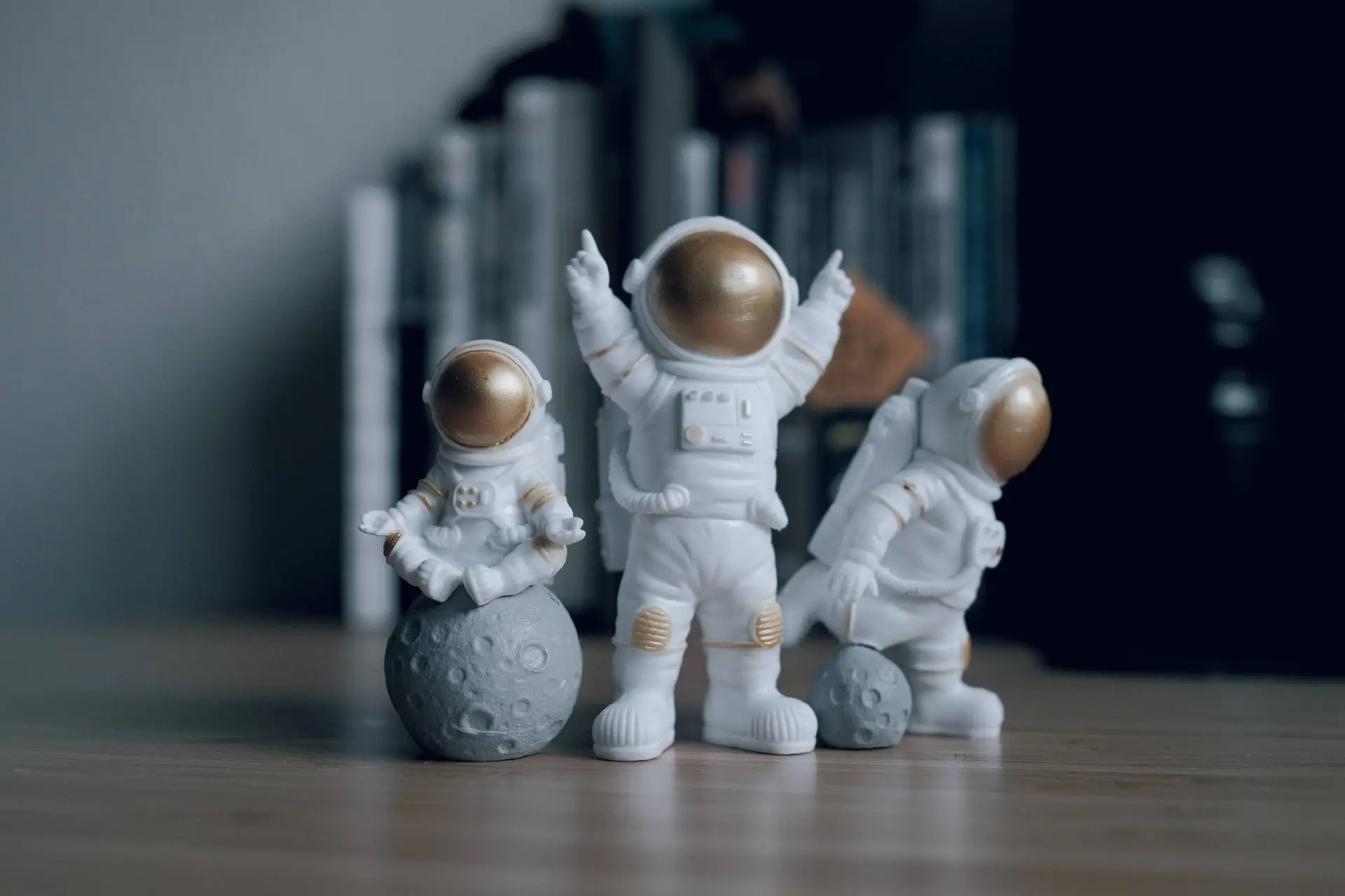 Three astronaut figurines are placed on a wooden surface. The smallest sits on a gray moon, the medium one stands with arms raised, and the largest kneels beside a small moon, evoking an imaginative world akin to space-themed food adventures. Books are blurred in the background.