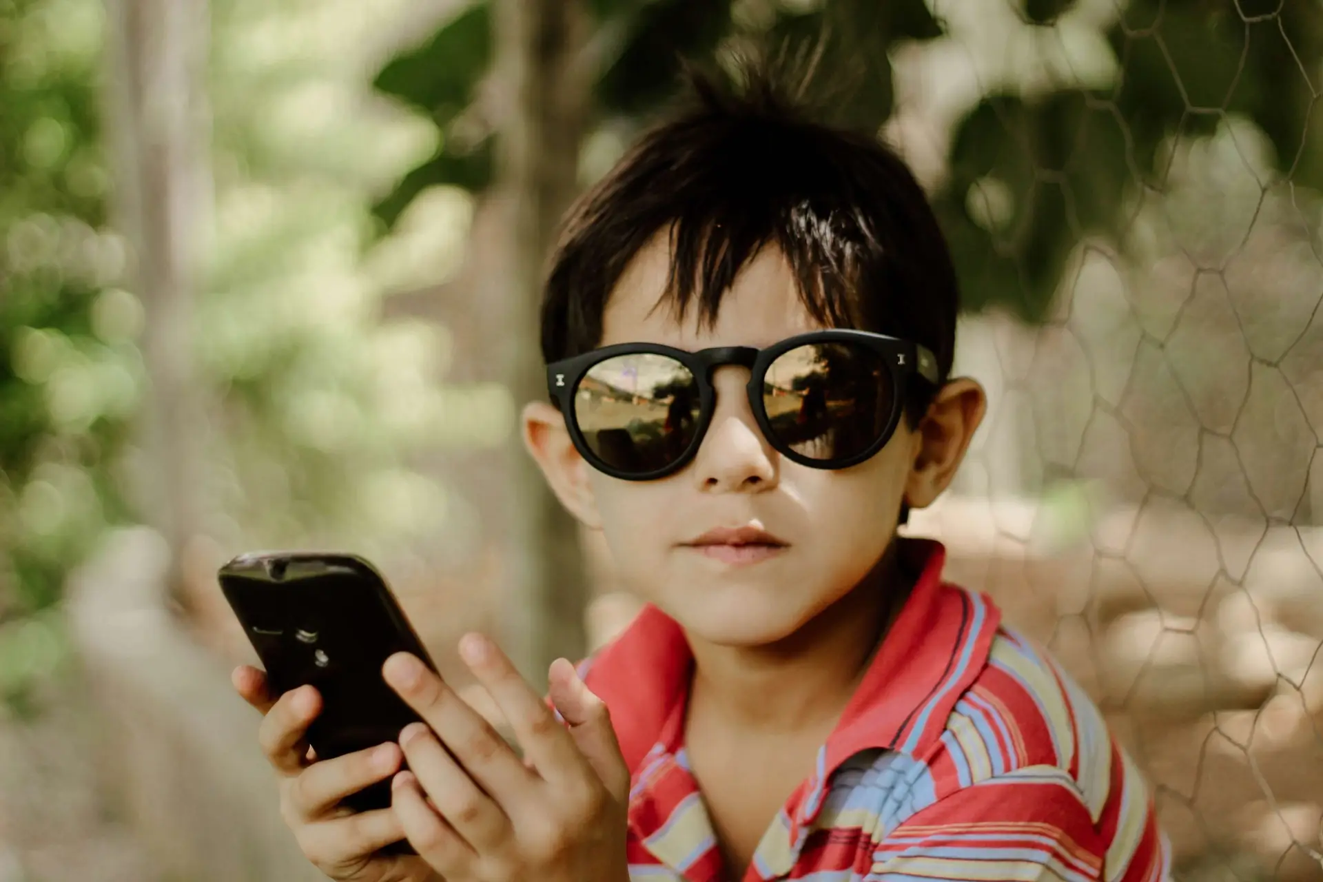 A young boy wearing sunglasses and a striped shirt holds a smartphone while sitting outdoors, watching engaging educational safety videos for kids. The background features greenery and a fence.