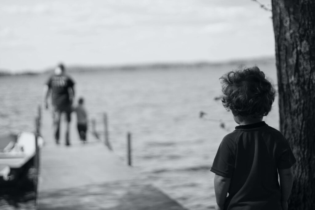 A black and white image captures a poignant moment: a child stands by a tree, gazing at the dock where two figures, an adult and another child, walk toward the water. Perhaps they're engaging in grief activities for kids as they connect with nature. A small boat is moored beside them.