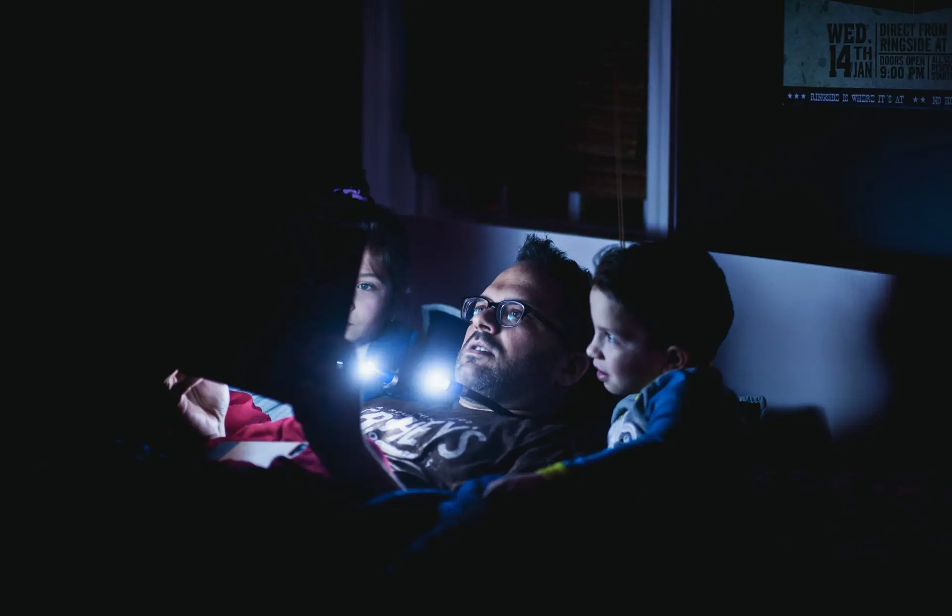 A man and two children lie in bed in a dark room, illuminated by a small light source. The man and children are focused on the light, discovering how to help kids with nightmares feel safe and comforted during the night.