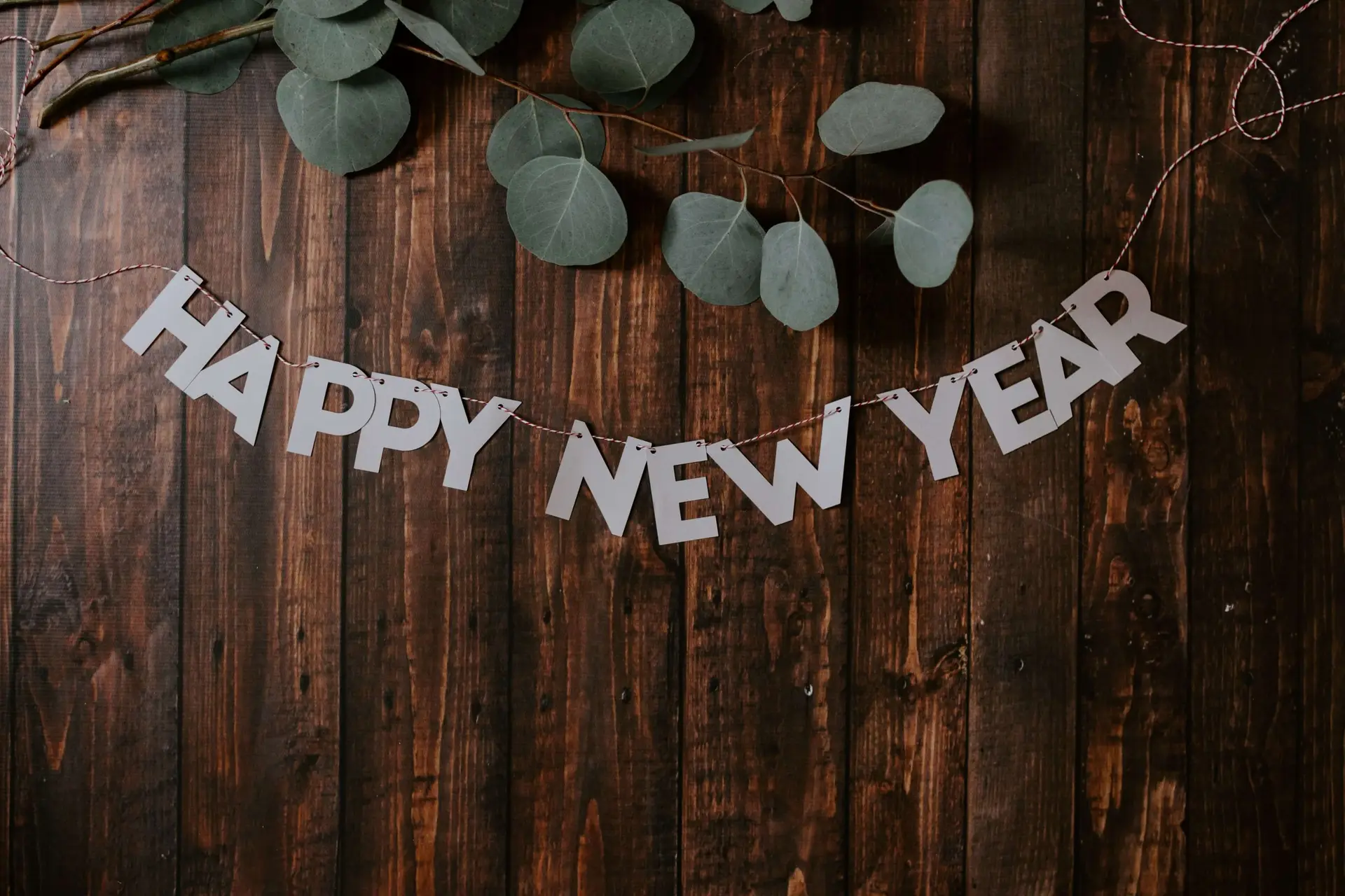 A HAPPY NEW YEAR banner hangs on a dark wooden surface, elegantly decorated with eucalyptus branches for a festive and natural touch, setting the perfect scene for a kids' New Year's Eve party.