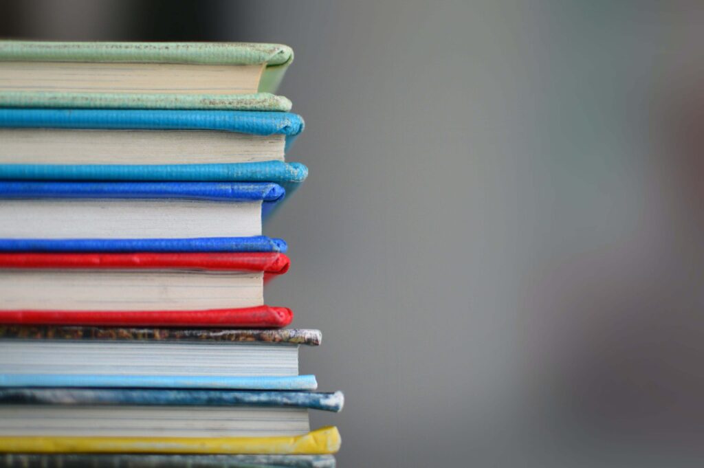 A stack of books with colorful covers, including titles like "The Babysitters Club," is piled on the left side of the image against a plain, blurred background. The spines display various colors such as blue, red, and green.