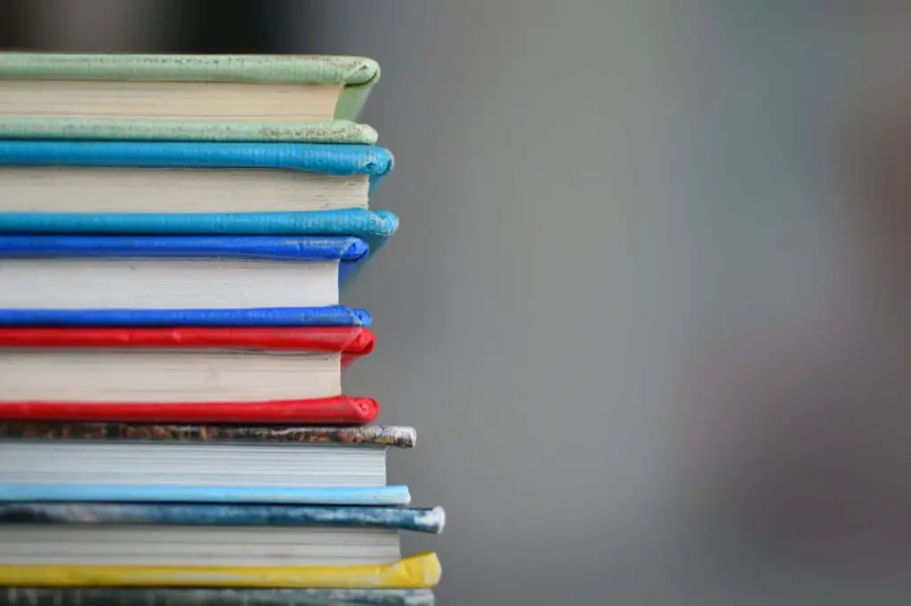 A stack of books with colorful covers, including titles like "The Babysitters Club," is piled on the left side of the image against a plain, blurred background. The spines display various colors such as blue, red, and green.