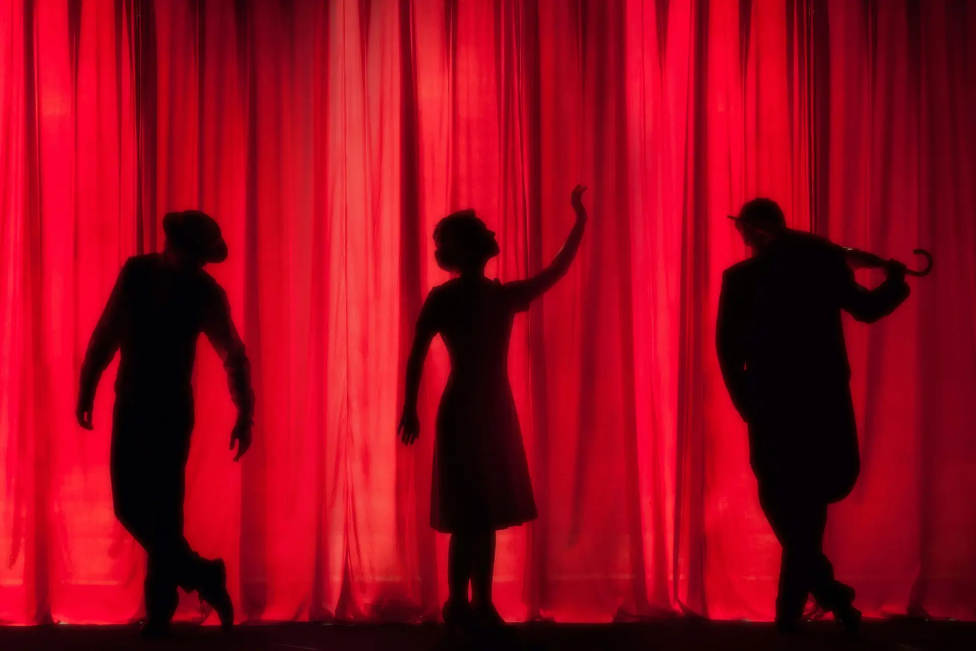 Silhouettes of three people striking dynamic poses in front of dramatic red curtains evoke the playful spontaneity of improv games for kids. One holds an umbrella, another raises a hand, and the third stands with hands on hips, creating a theatrical and artistic scene.