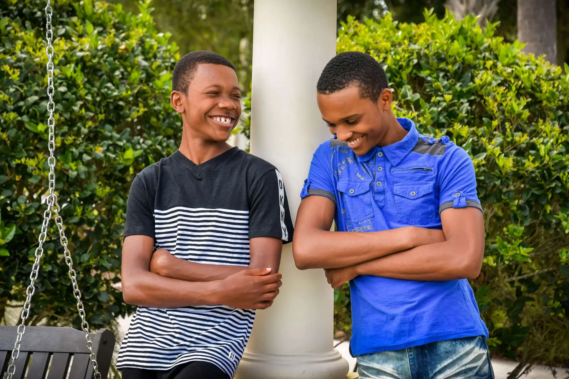 Two young boys, sharing summer jokes for kids, are standing outside, laughing and leaning against a white pillar. One boy wears a black-and-white striped shirt, while the other sports a blue shirt. Lush greenery surrounds them in the background.