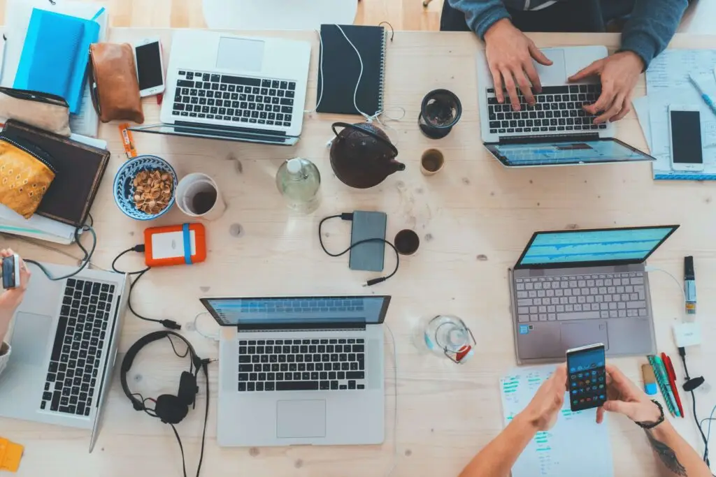 Aerial view of a cluttered desk with multiple laptops, a tablet, notebooks, headphones, snacks, and a teapot. Four people are engaged in work, typing and discussing child predators on social media while using a smartphone. Various personal items and papers are scattered across the workspace.