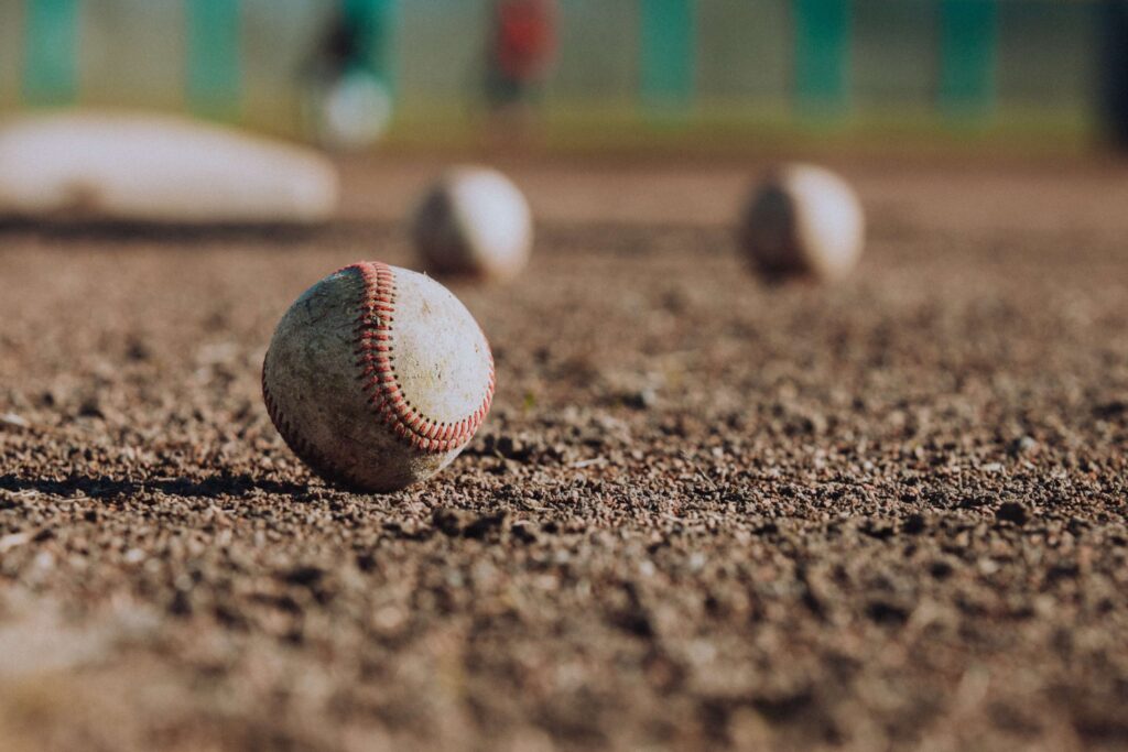 Close-up of a worn baseball on a dirt field, reminiscent of classic kids' baseball movies, with other blurred baseballs and a base visible in the background. The scene suggests a sunny day at a bustling baseball diamond.