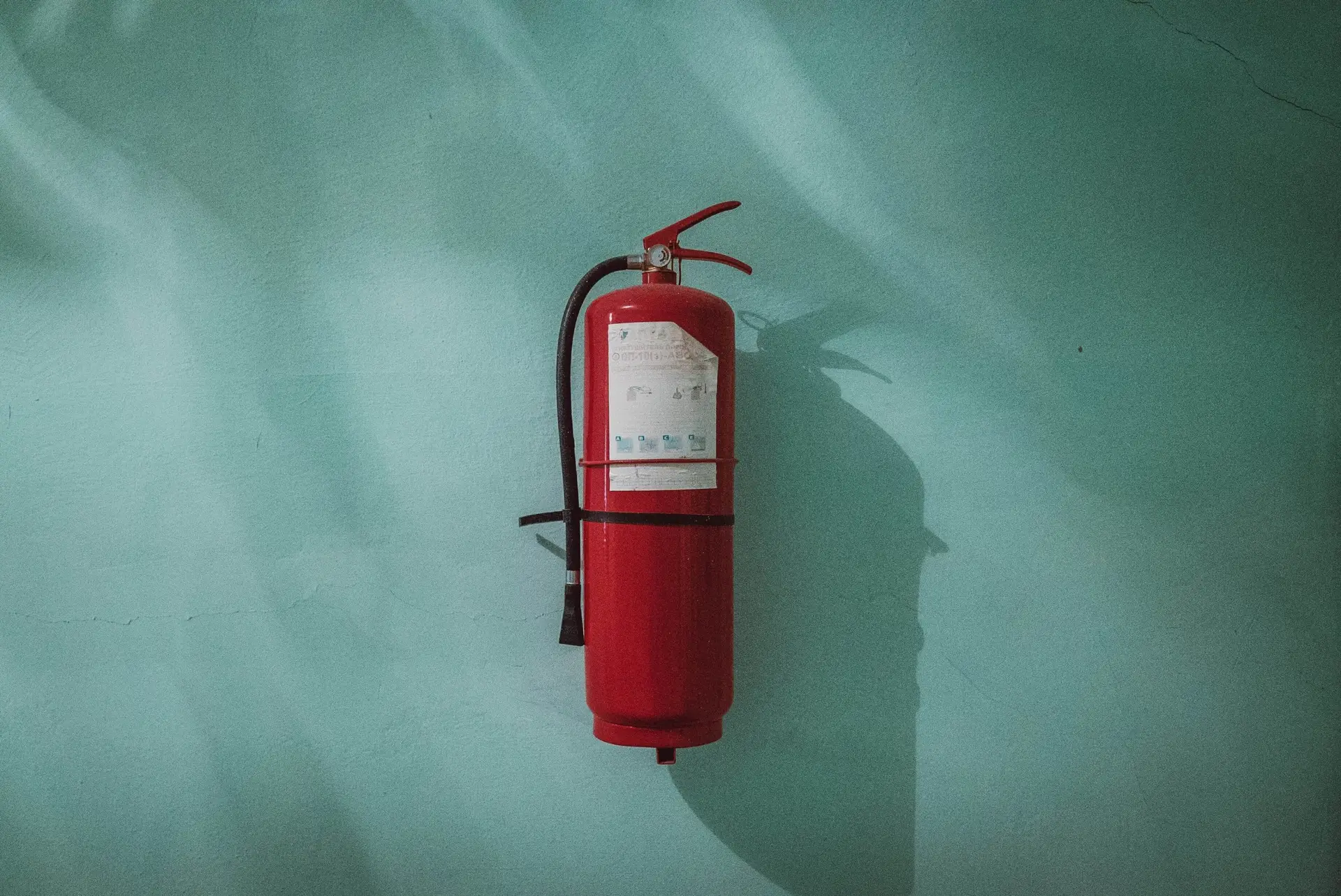 A red fire extinguisher mounted on a light teal wall sparks curiosity, inviting safety questions for kids. Shadows dance, highlighting the wall's texture and cracks. While the extinguisher's instructions are visible, they remain unreadable from this angle.