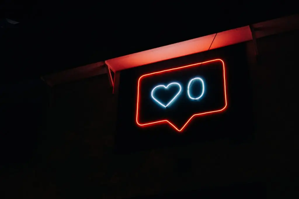 A neon sign shaped like a social media notification bubble glows against the dark background, echoing real-life negative social media stories. It features a heart symbol followed by the number zero, outlined in red, with the designs illuminated in blue.