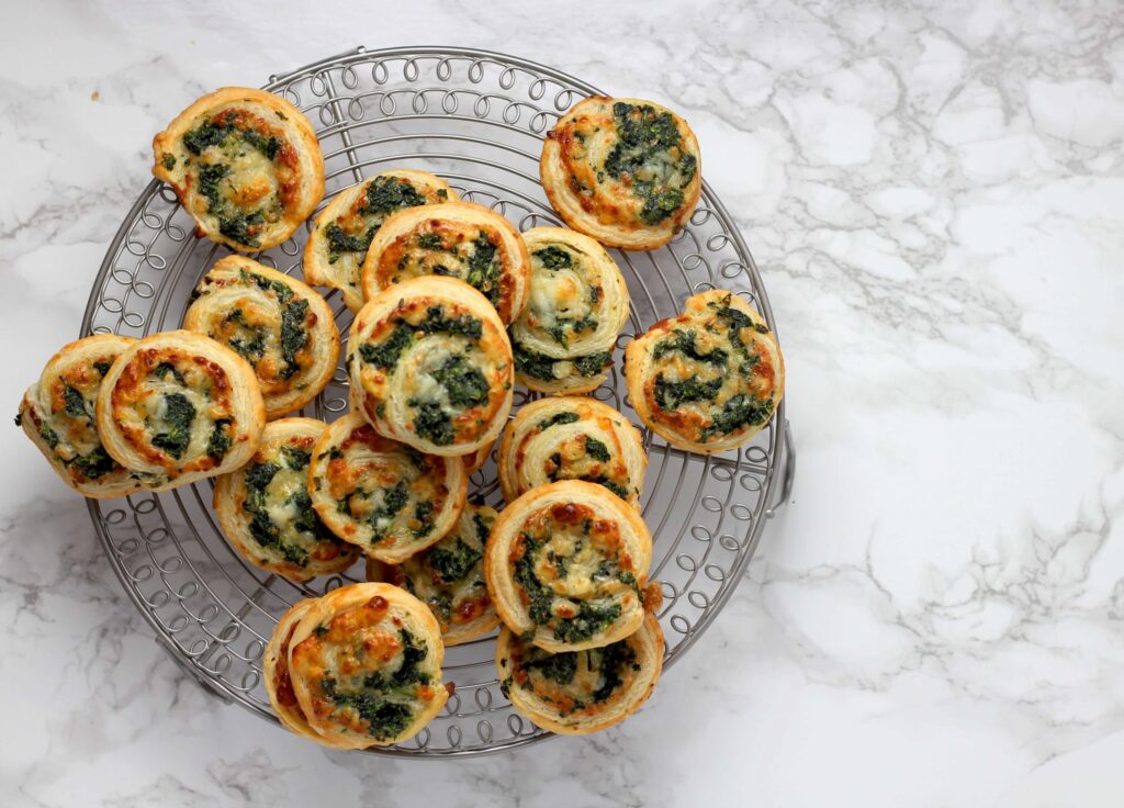 A wire rack on a marble surface holds freshly baked, kid-friendly appetizers—savory pastries with swirls of spinach and cheese. The golden-brown pastries boast melted cheese and green spinach flecks, making them perfect for little ones.