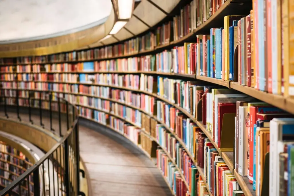 A curved library shelf filled with colorful, realistic fiction books for kids stretches into the distance. A black railing runs parallel to the shelves, and soft light illuminates the scene, creating a warm, inviting atmosphere.