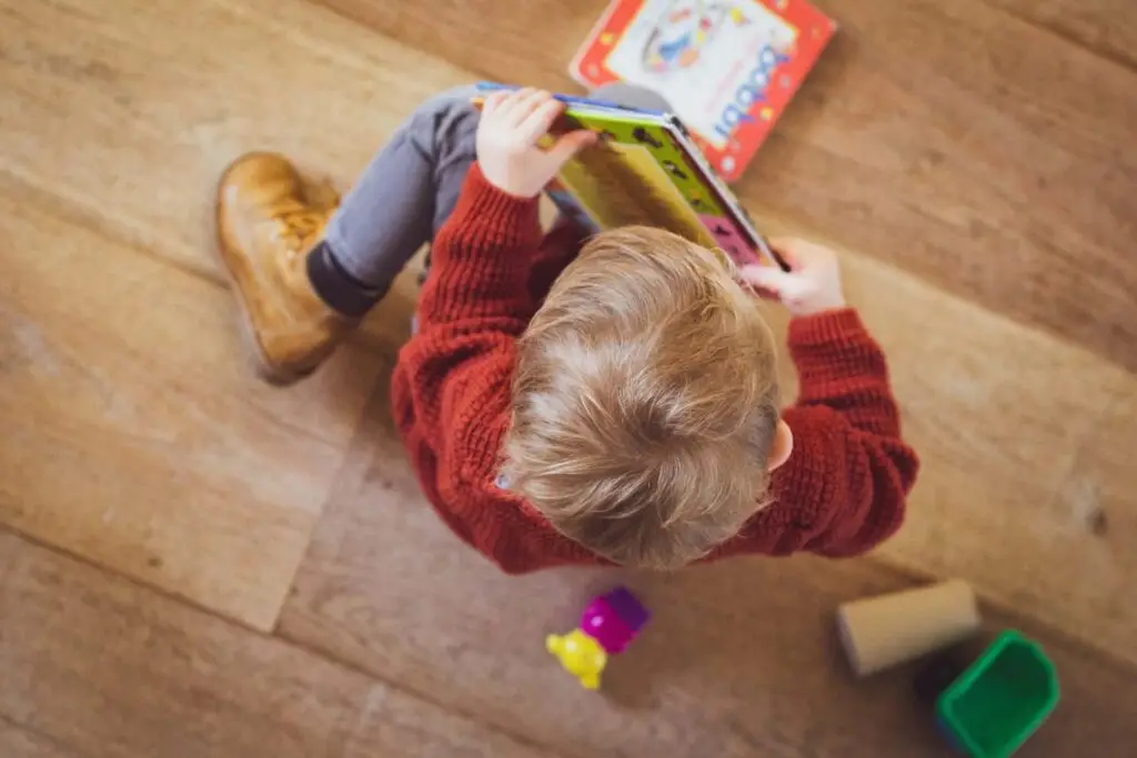 A young child with blonde hair, wearing a red sweater and brown boots, sits on a wooden floor engrossed in an open book. Colorful toys and a small cup are scattered nearby, sparking curiosity about the world beyond even inappropriate children's books.