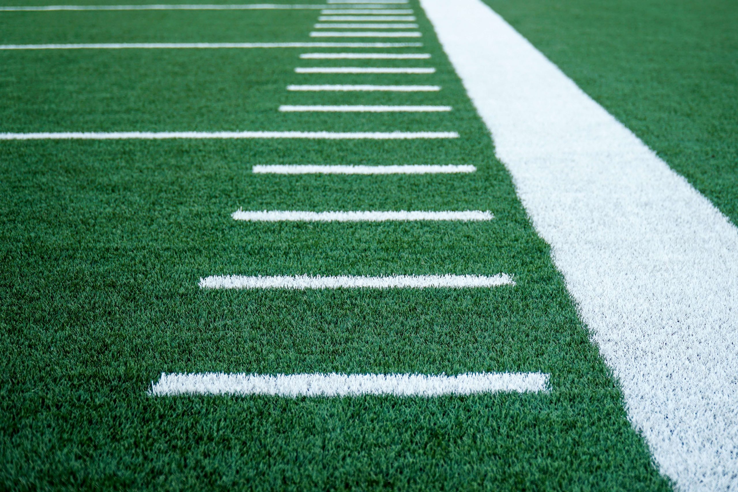 Close-up view of a football field showing green artificial turf with white yard lines and a wide sideline on the right, reminiscent of the miniature fields found in football toys for kids. The lines create a pattern extending into the distance, indicating yard markers on the field.