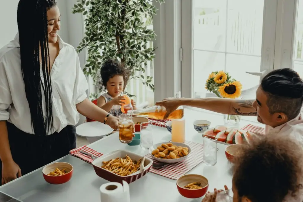 A family of four sharing a meal at a dining table adorned with chips, fruit, and a vase of sunflowers. Amidst the laughter and conversation, when to talk to kids about sex is thoughtfully considered as one person pours a drink while another serves a child.