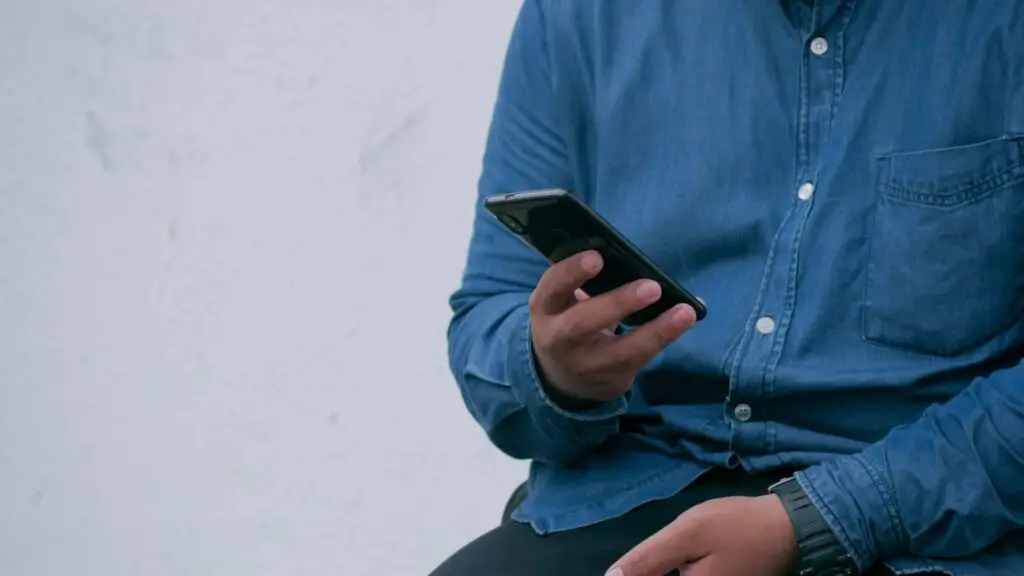 Person in a blue denim shirt holding a smartphone focuses intently on the screen, seated against a plain white background. As they scroll, one might wonder what causes cyberbullying and how it impacts online interactions, drawing attention to the importance of digital awareness.