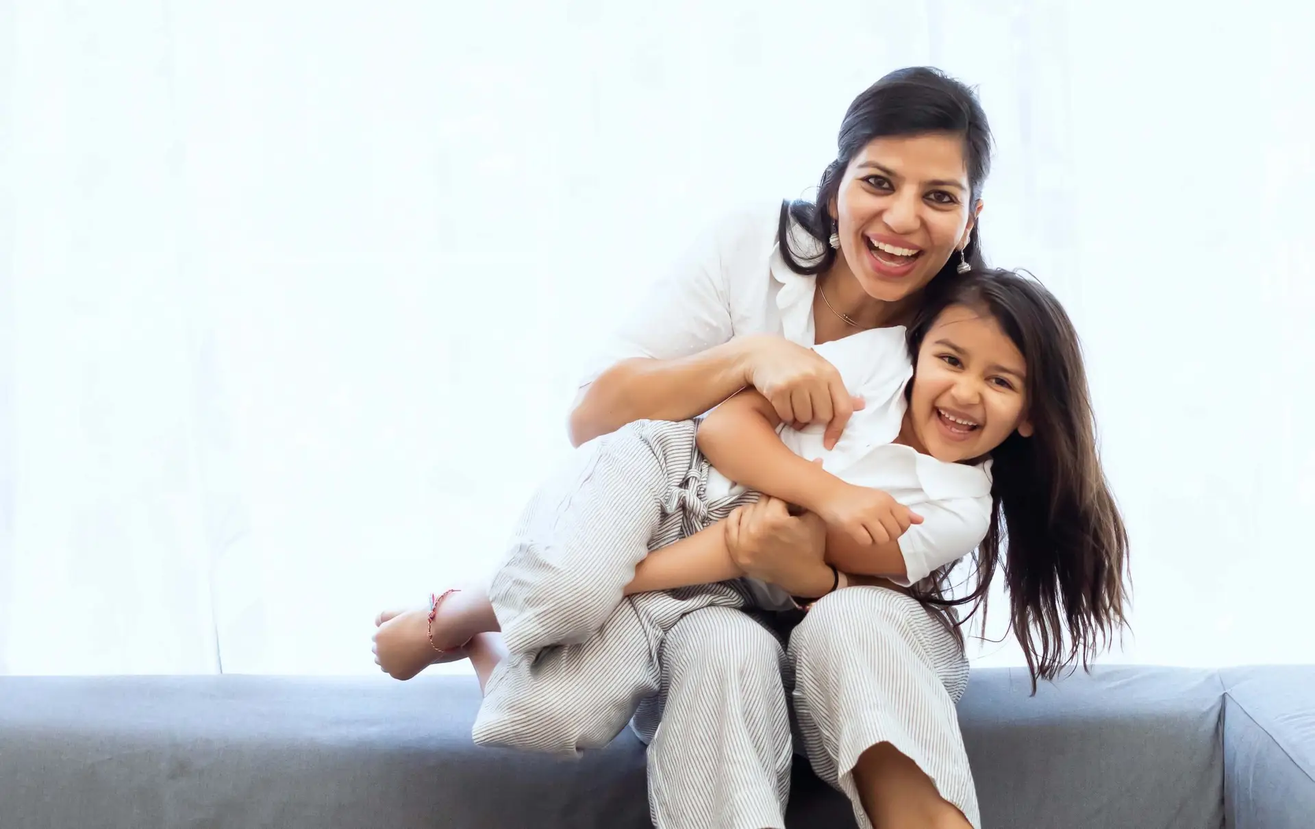 A woman and a young girl are sitting on a sofa, laughing and playfully embracing. Both are wearing light-colored clothing, and the softly lit background creates a warm atmosphere, underscoring the importance of safety and knowing how to report a child predator to protect such joyful moments.