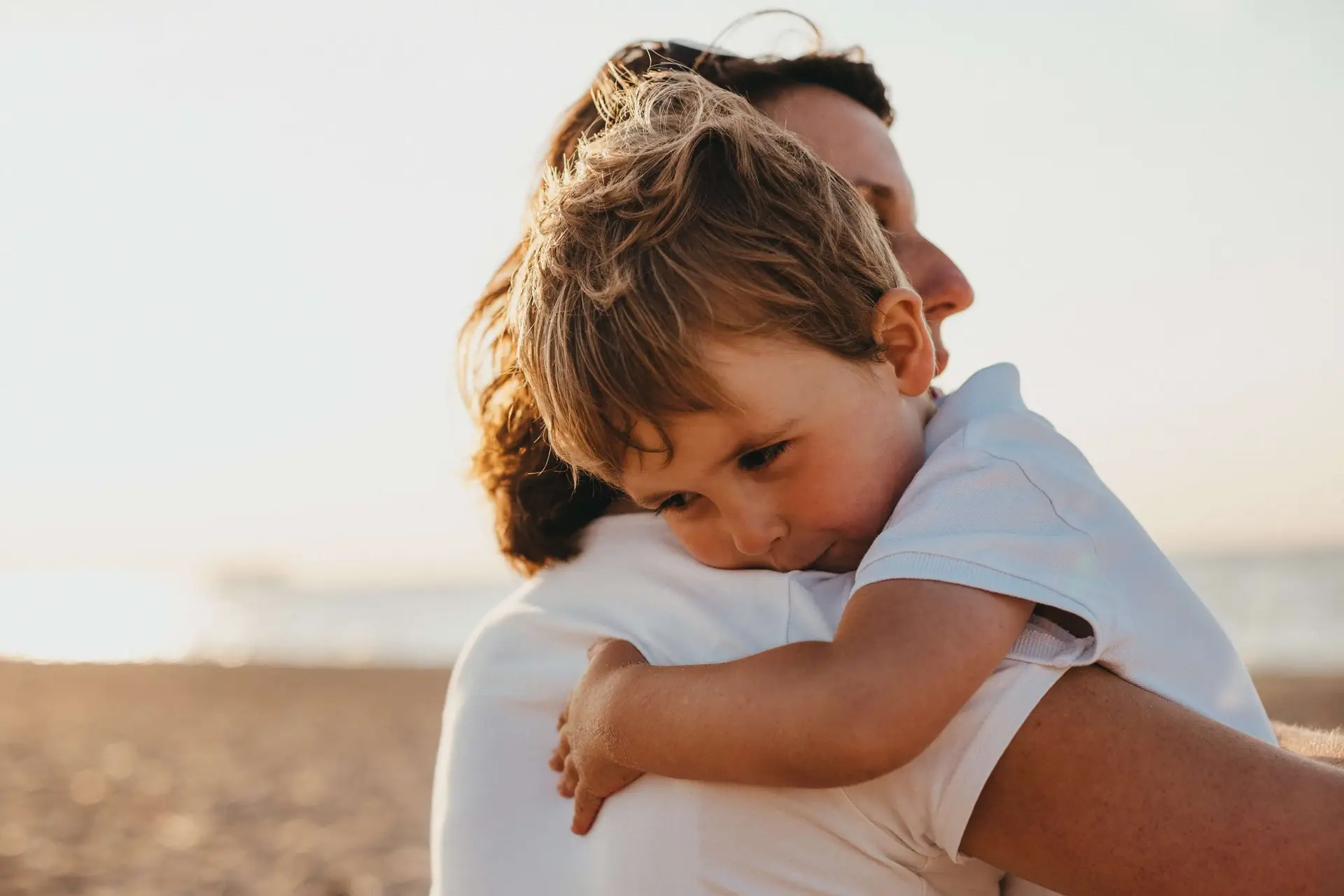 A young child with short, light hair is being warmly embraced by an adult at the beach, embodying the spirit of empathy activities for kids. The child looks thoughtful in a white shirt, with sand and a soft-focus view of the ocean and sky in the background.