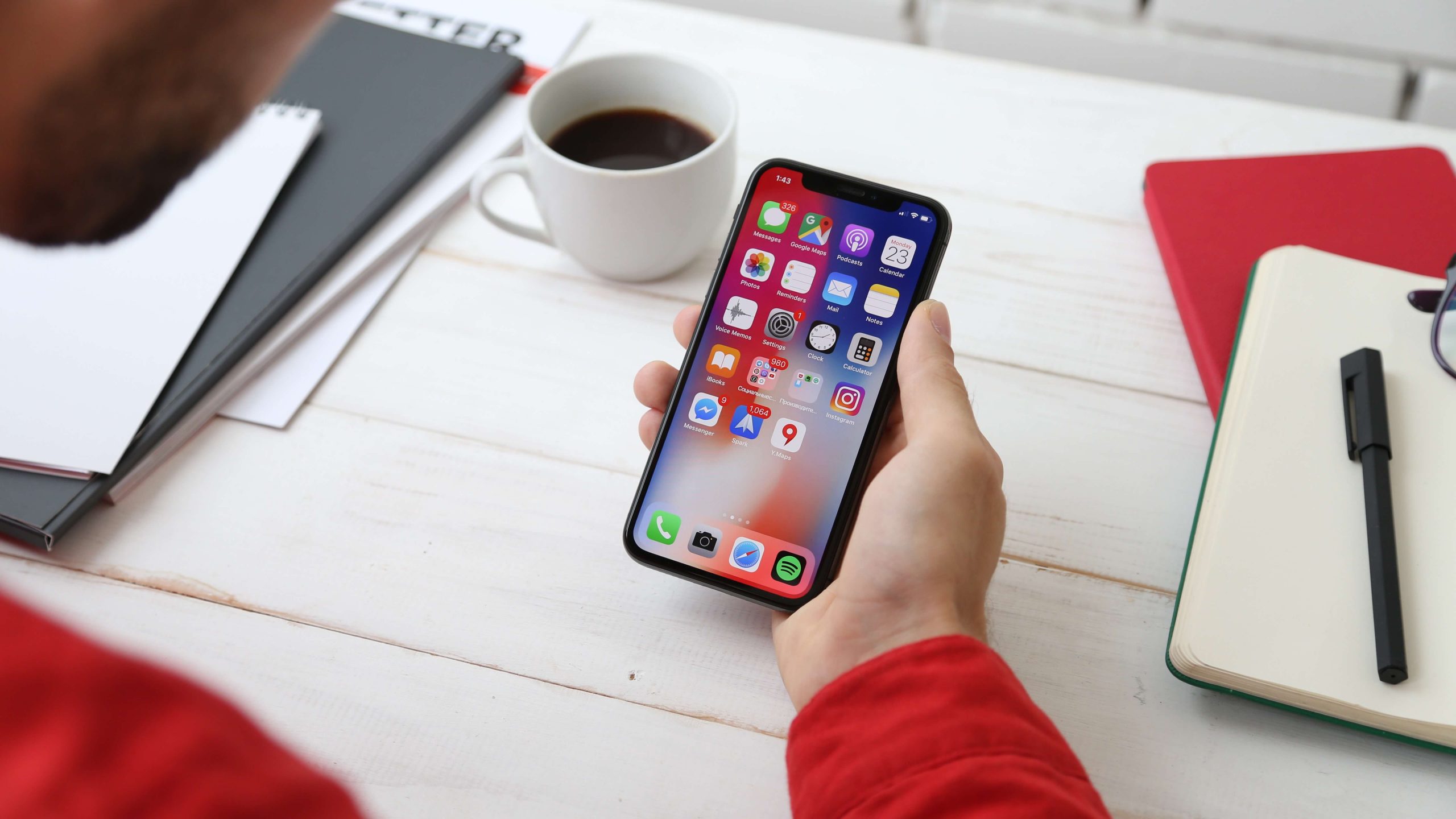 A person wearing a red shirt holds a smartphone displaying various app icons, including one designed to keep kids safe. The phone is held over a white desk with a cup of coffee, notebooks, and a pen.