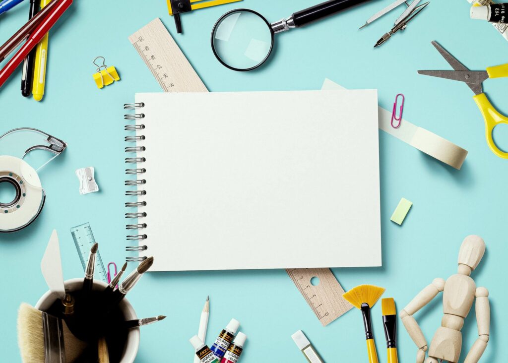 A blank spiral notebook, perfect for your back-to-school checklist, is surrounded by an array of art and office supplies including pencils, scissors, a ruler, paints brushes, paper clips, and a wooden mannequin—all set against a light blue background.