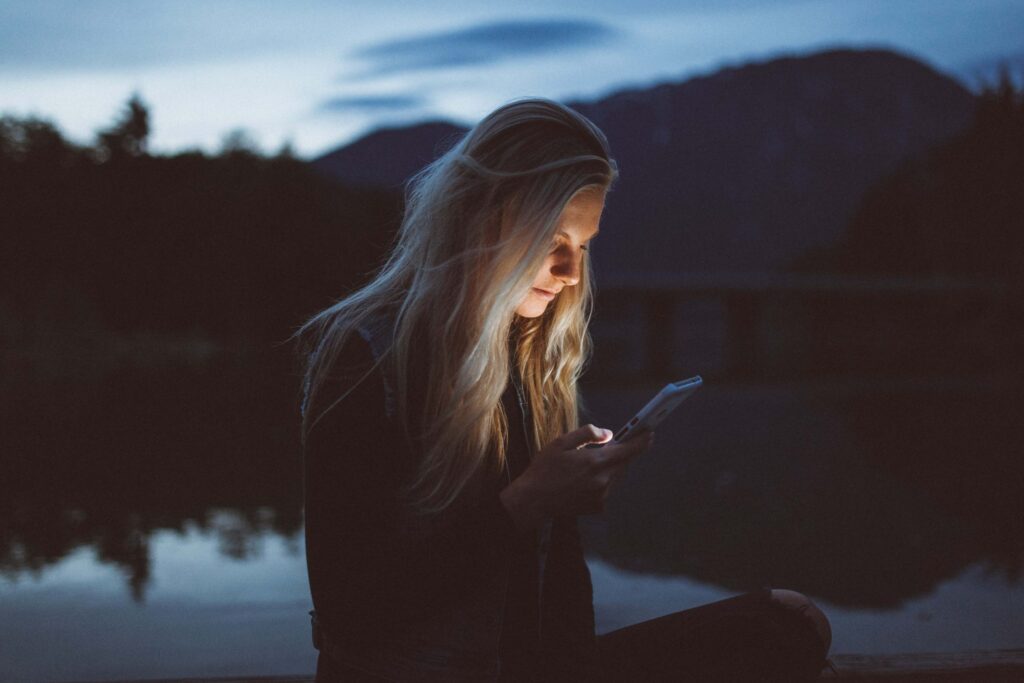 A person with long hair sits outdoors by a body of water at dusk, pondering if social media affects social skills while looking at a smartphone. The background features a silhouette of trees and mountains against a softly dimly lit sky.