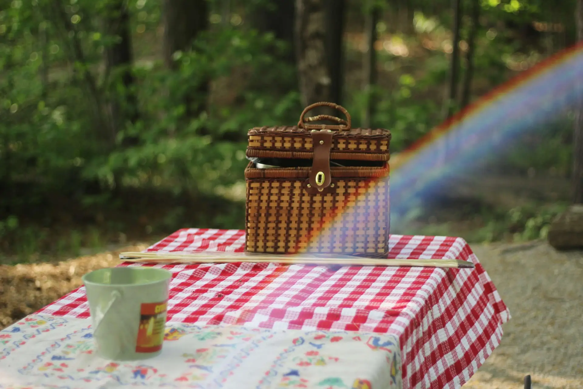 A wicker basket sits on a red-and-white checkered tablecloth at a quaint kids' picnic table, with a colorful rainbow arc crossing over it. Nearby, a green mug rests on an embroidered cloth, surrounded by a lush green forest background, painting a picture-perfect outdoor scene.